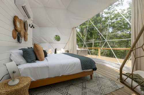 Cozy interior of a geodesic dome with a bed, natural decor, and large windows overlooking a forested area.