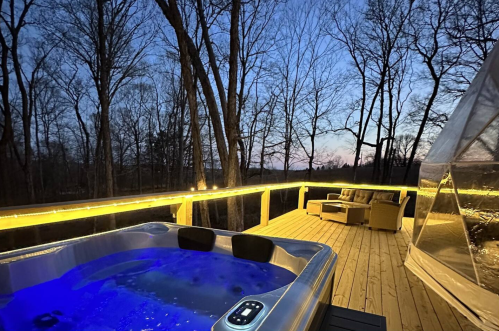 A hot tub with blue lighting on a wooden deck surrounded by trees at dusk, with a cozy seating area nearby.