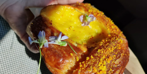 A hand holding a pastry topped with yellow filling and small flowers, resting on a textured surface.