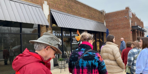 A line of people waiting outside a store with a sign that says "Give Good Bread" on a cloudy day.