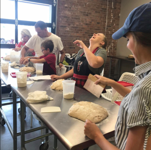 A group of people laughing and baking together at a table with dough and ingredients in a bright kitchen.
