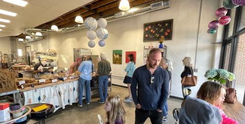 A busy art studio with people browsing artwork and displays, featuring colorful decorations and a warm atmosphere.