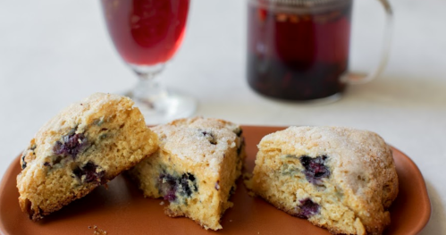 Three pieces of blueberry scone on a brown plate, with a glass of red drink and a mug of tea in the background.