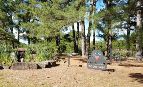 A peaceful outdoor area featuring a fountain, surrounded by trees and greenery, with informational signs nearby.