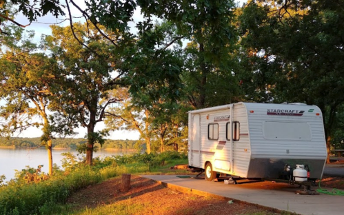 A white RV parked by a serene lake, surrounded by trees and bathed in warm sunlight.