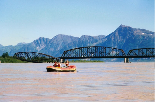 A small inflatable boat floats on a river with a mountain backdrop and a bridge in the distance under a clear blue sky.