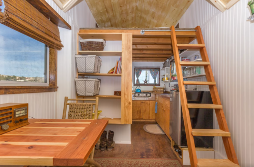 Cozy tiny house interior featuring a wooden loft, kitchen, and a small dining area with a ladder and natural light.