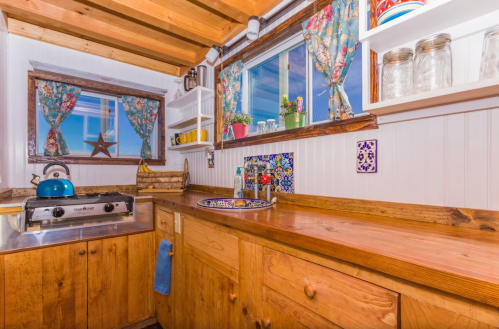 Cozy kitchen with wooden cabinets, colorful curtains, a blue kettle, and decorative tiles on the countertop.