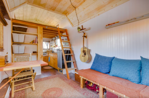 Cozy tiny home interior with a wooden loft, kitchen area, blue cushions, and an acoustic guitar on the wall.