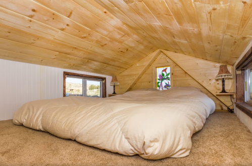 Cozy attic bedroom with a large bed, wooden ceiling, and two windows, featuring soft lighting and a warm atmosphere.