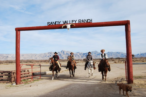 Four cowboys on horseback ride through the entrance of Sandy Valley Ranch, with mountains in the background. A dog walks nearby.