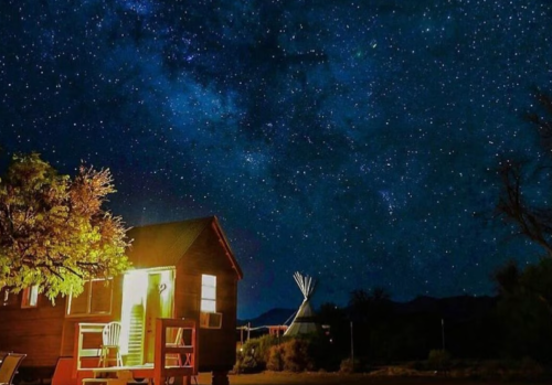 A cozy cabin and teepee under a starry night sky, with a warm glow from the cabin's window.