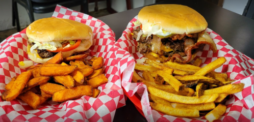 Two burgers with toppings on red checkered paper, accompanied by a side of sweet potato and regular fries.