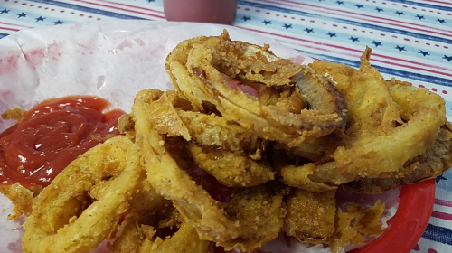 A plate of crispy, golden-brown onion rings served with a side of ketchup on a red and white checkered tablecloth.