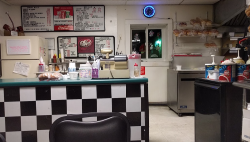 A diner interior featuring a checkered counter, menu board, condiments, and a display of baked goods.