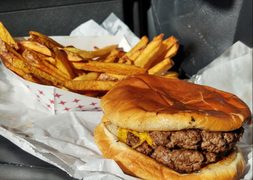 A close-up of a double burger with melted cheese, served with a side of golden French fries in a takeout container.