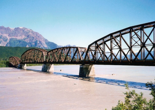 A rusted metal bridge spans a wide river, with mountains in the background under a clear blue sky.