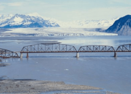 A rusted metal bridge spans a river with snow-capped mountains and a glacier in the background.
