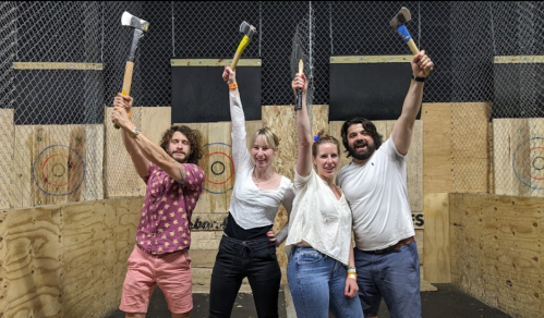 Four friends pose with raised axes in an axe-throwing venue, smiling and celebrating their fun experience.