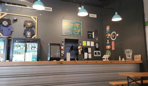 A cozy bar with a wooden counter, colorful wall decor, and a display of drinks in a fridge.