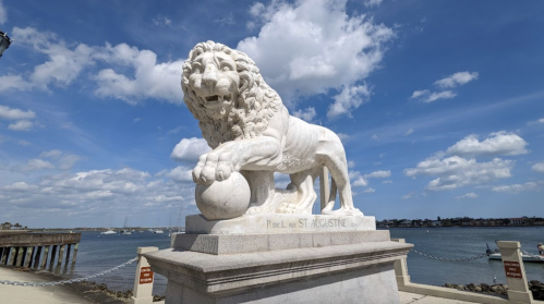 A white lion statue holding a ball, overlooking a waterfront with boats and a cloudy sky in the background.