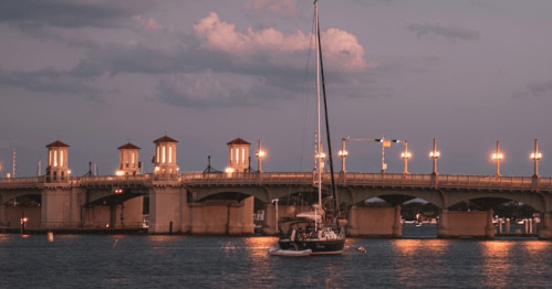 A sailboat anchored near a lit bridge at dusk, with clouds and a calm water surface reflecting the lights.