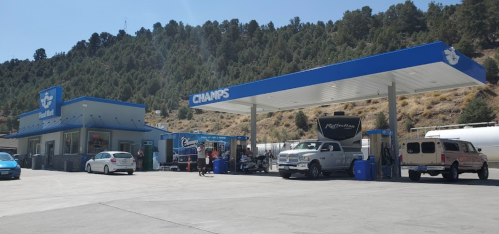 A gas station with a blue canopy, cars refueling, and a mountainous backdrop under a clear sky.