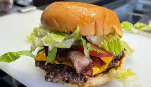 A close-up of a juicy burger with lettuce, tomato, cheese, and bacon on a toasted bun, resting on a white cutting board.