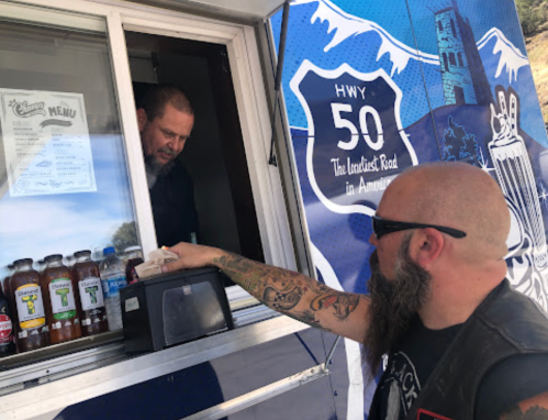A customer hands cash to a vendor at a food truck, with a scenic mountain backdrop and a menu displayed.