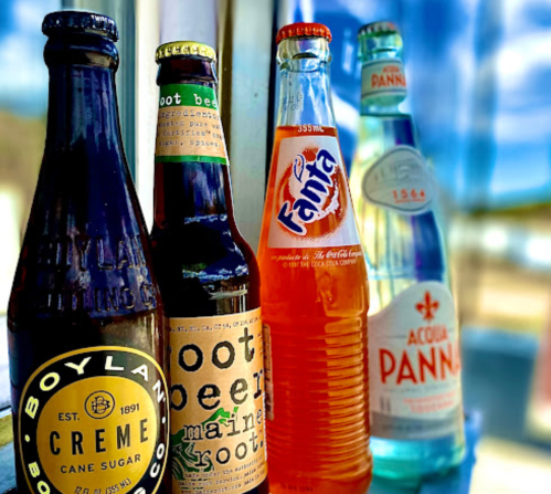 Four colorful glass bottles of beverages: Boylan Creme, Maine Root Beer, Fanta, and Acqua Panna, against a bright background.