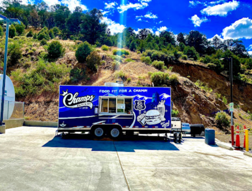 A food truck named "Champs" parked in a scenic area with trees and hills in the background.