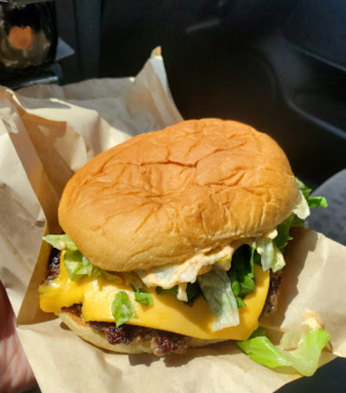 A hand holding a cheeseburger with lettuce, sauce, and a soft bun, resting on a piece of brown paper.