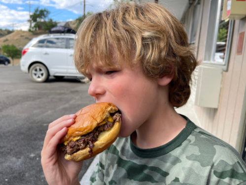 A young boy with curly hair prepares to take a big bite of a juicy hamburger outside.