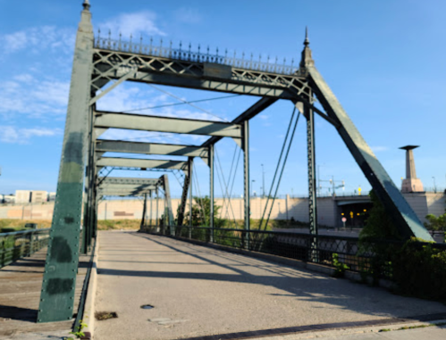 A historic metal bridge with a blue sky and greenery, featuring intricate designs and a clear pathway.