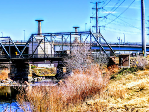A metal bridge spans a river, with power lines and a grassy bank in the background under a clear blue sky.