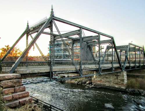 A metal bridge spans a river, with modern buildings in the background and a sunset casting warm light.