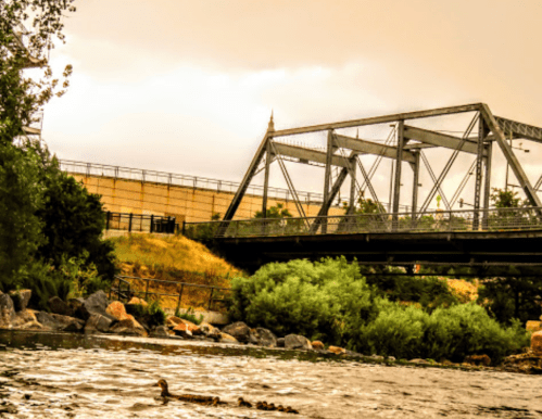 A metal bridge spans a river, surrounded by greenery and rocky banks under a cloudy sky.