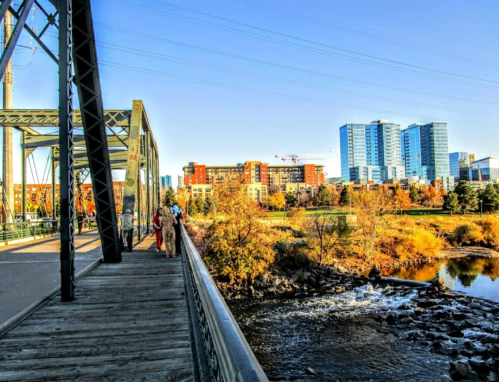 A scenic view of a bridge with people walking, overlooking a river and modern buildings in the background.