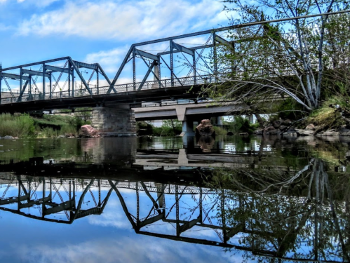A metal bridge spans a calm river, reflecting its structure and surrounding greenery under a blue sky.