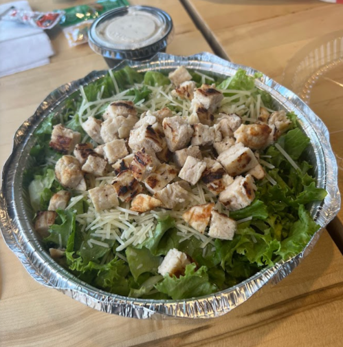 A salad in a foil container, topped with grilled chicken pieces and shredded cheese on a bed of leafy greens.
