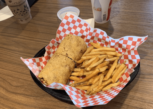A wrap sandwich on a checkered paper plate, accompanied by a side of French fries and a drink.