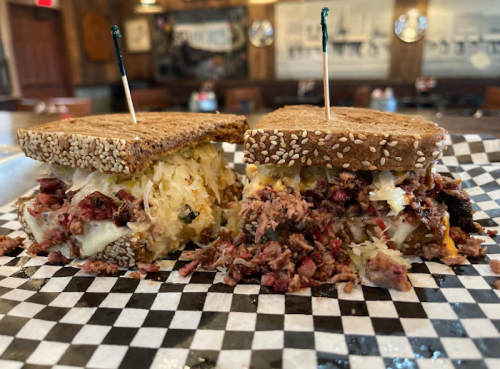 A close-up of a sandwich with layers of meat, sauerkraut, and cheese on sesame seed bread, served on a checkered paper.