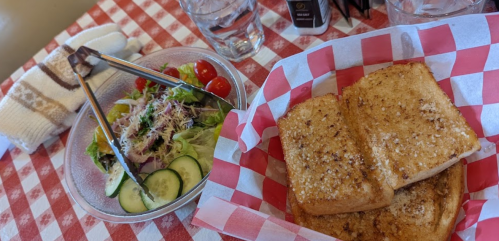 A basket of toasted bread with a side salad of greens, cucumbers, and cherry tomatoes on a checkered tablecloth.