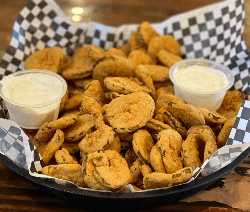 A basket of crispy fried pickles served with two cups of dipping sauce on a checkered paper liner.