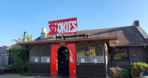 A rustic restaurant with a red sign reading "Smokies" and an open sign in the window, surrounded by greenery.