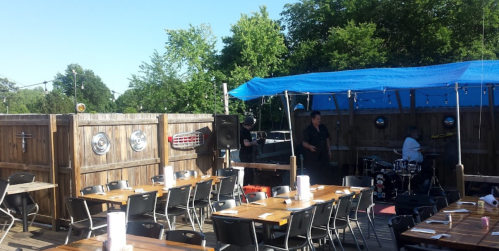 Outdoor dining area with wooden tables, a blue canopy, and a small stage for live music. Trees in the background.