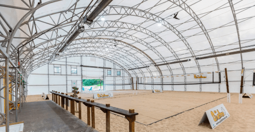 Indoor sand volleyball court with a large, arched structure and sandy floor, featuring volleyball nets and signage.