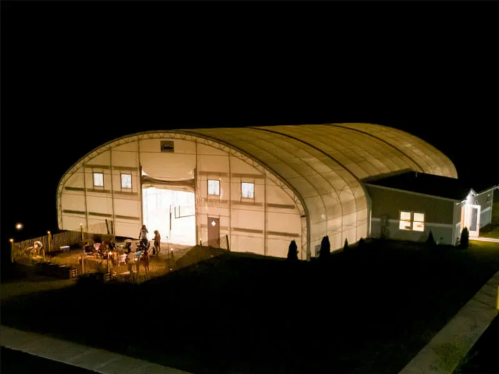 A large, illuminated indoor structure at night, with people gathered outside on a patio.