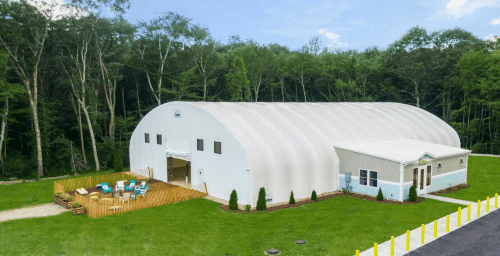 A large white dome-shaped building with a deck and seating area, surrounded by green grass and trees.