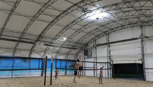 A group of children playing volleyball indoors on a sandy court under a large, arched tent structure.